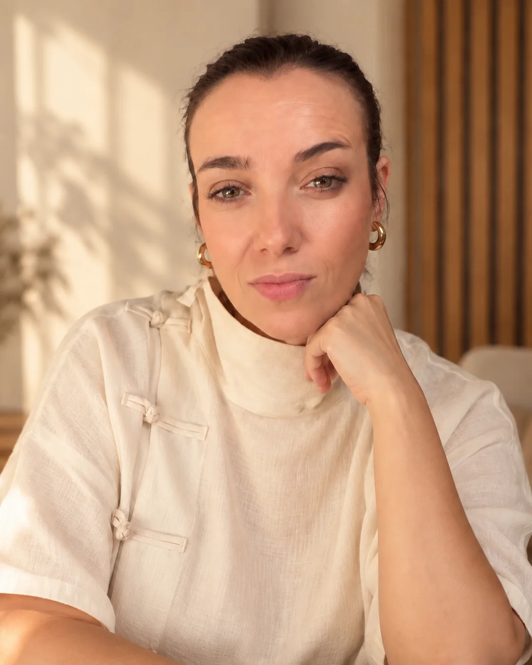 Vanesa Viciano, psicóloga, sentada con la mano bajo la barbilla, con una blusa de lino crema y luz cálida de tarde